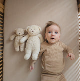 Moonie Sensorik-Teddybär im Farbton Organic Sand neben Einschlafbär und Kleinkind in Bett liegend
