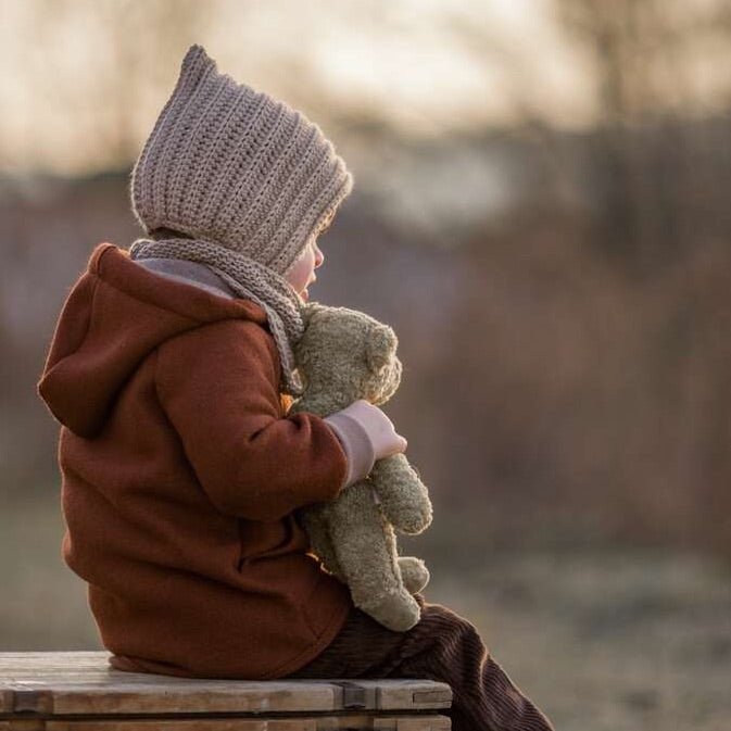 Kind sitzt draußen mit Schlenker Kuscheltier kleiner Bär in Beige von Senger Naturwelt