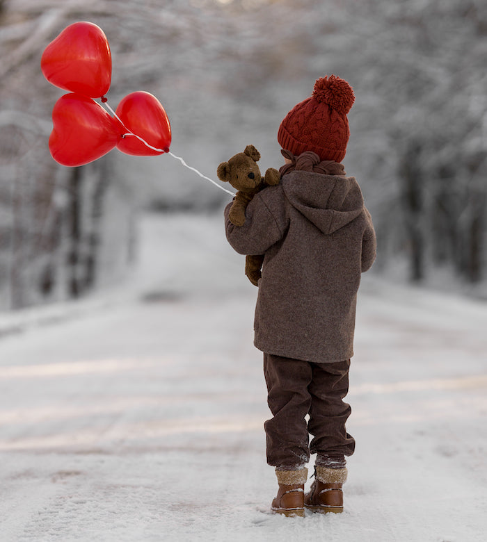 Kind mit Braunem Bär Schlenker Kuscheltier von Senger Naturwelt sowie roten Luftballons