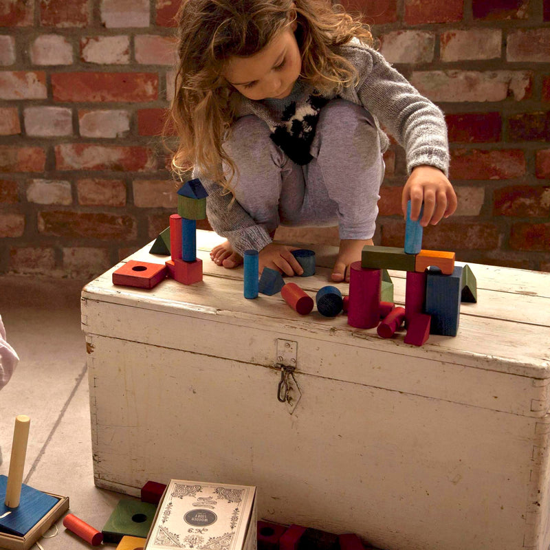 Kind auf Holzkiste am Spielen mit 30-teiligem Set an Holzbauklötzen in Regenbogenfarbe von Wooden Story